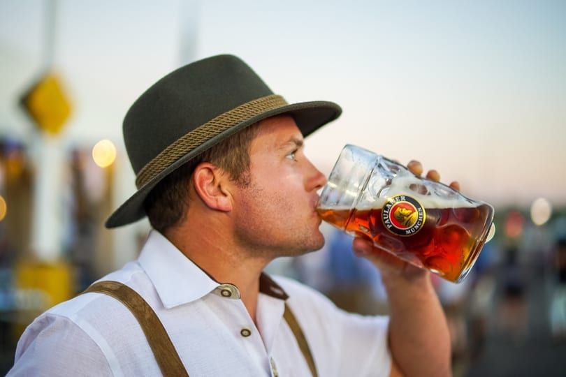 Man in a hat and suspenders drinking from a large glass of beer.