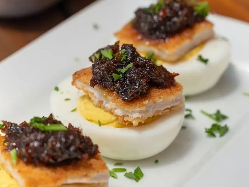 Three deviled eggs topped with fried chicken and dark relish, garnished with parsley on a white plate.