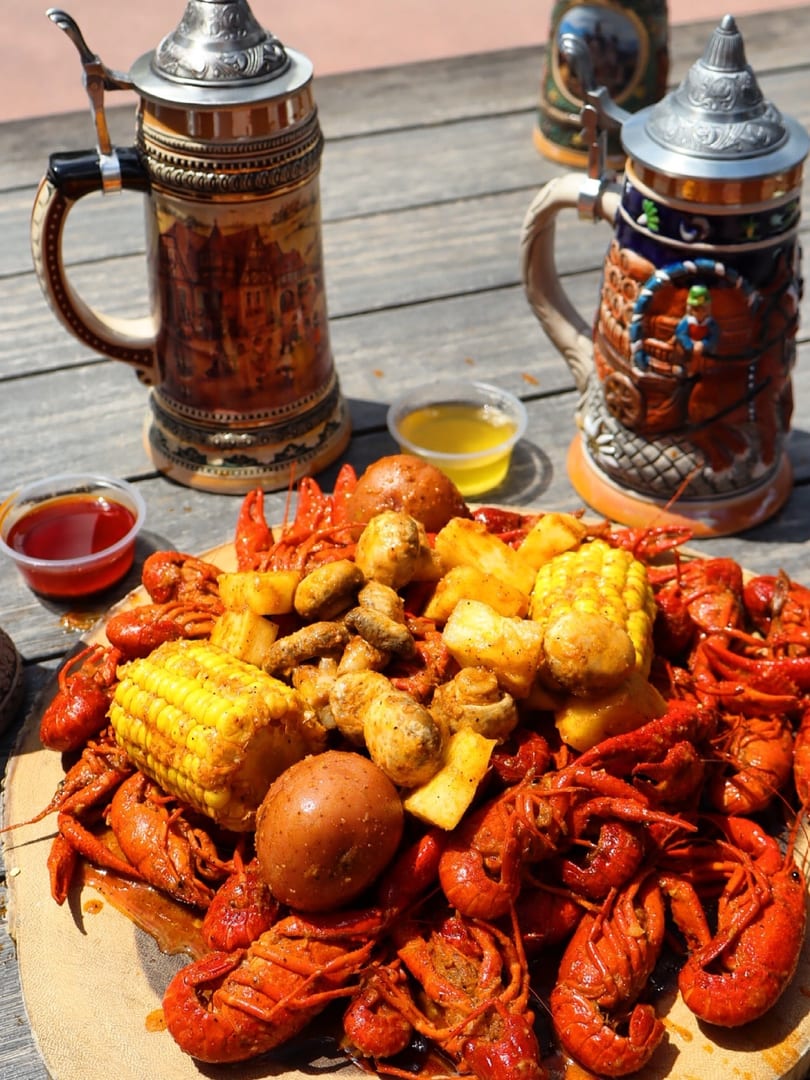Boiled crawfish, corn, potatoes, and mushrooms on a wooden board with two beer steins.