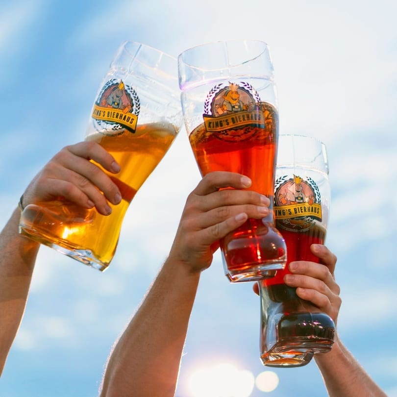 Three hands hold up King's Bierhaus branded beer glasses, filled with different colored beers, against a blue sky.