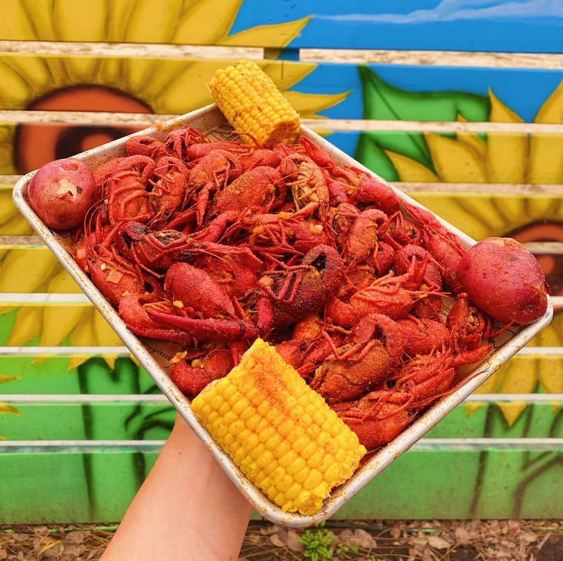 A tray of seasoned red crawfish, corn on the cob, and potatoes held against a sunflower mural.