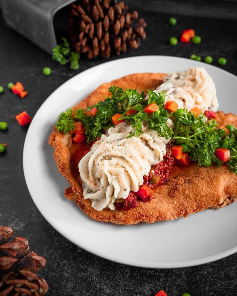 Crispy breaded cutlet topped with mashed potatoes, red sauce, peas, carrots, and parsley on a white plate.
