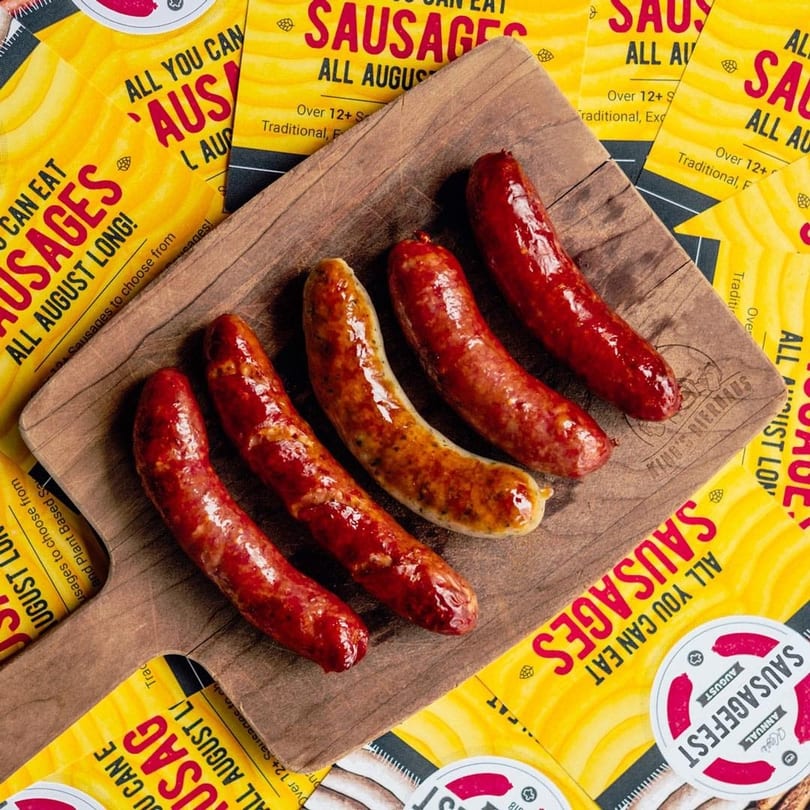 Five grilled sausages, some red and some golden brown, on a wooden board amidst yellow promotional flyers.