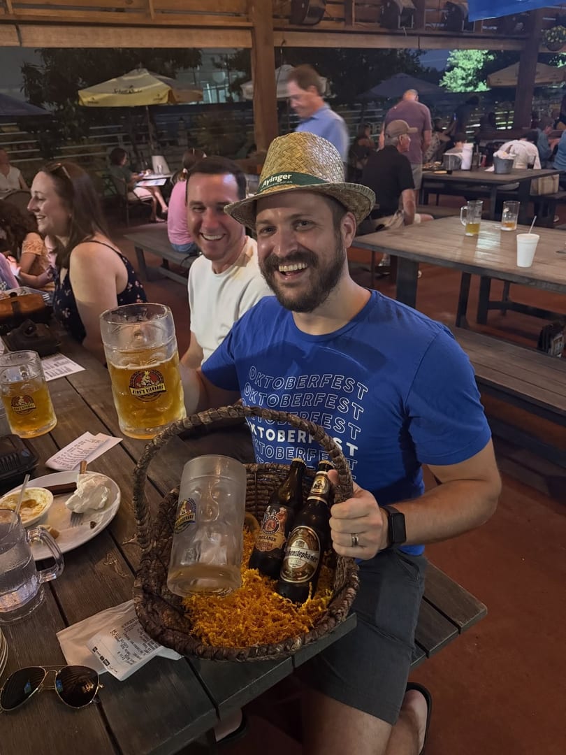 Man in Oktoberfest shirt and straw hat holding a beer basket, smiling at a lively outdoor table.