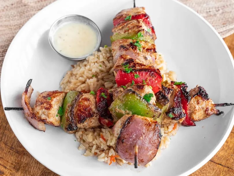 Two chicken and vegetable skewers with rice and a side of sauce on a white plate.
