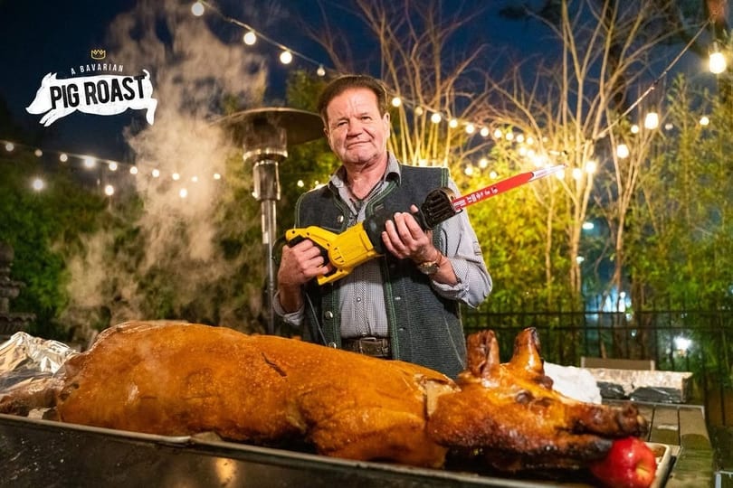 Man in Bavarian attire holding a saw next to a roasted pig with an apple in its mouth.