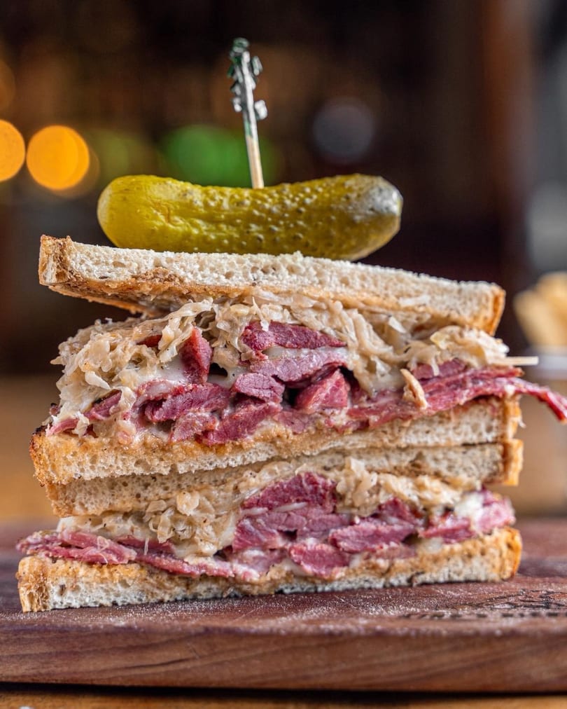 Stacked pastrami and sauerkraut sandwich on toasted bread, with a pickle spear on top.