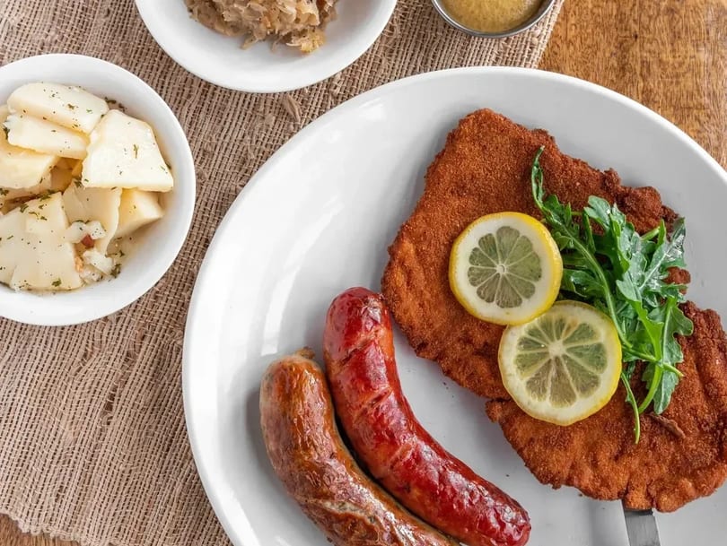 A plate of schnitzel with lemon slices and arugula, two sausages, and sides of potato salad and sauerkraut.
