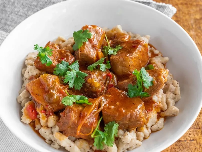 Hearty goulash with tender meat and dumplings, garnished with fresh cilantro in a white bowl.