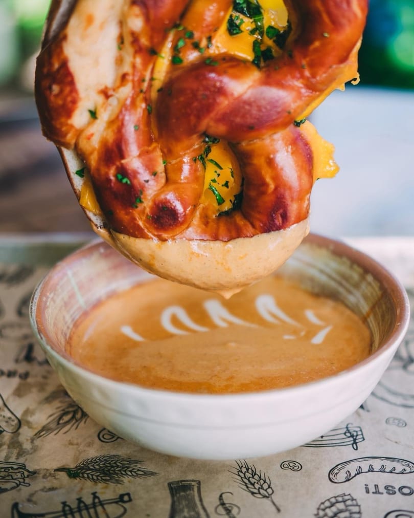 A cheesy pretzel being dipped into a bowl of creamy orange soup with white swirls.